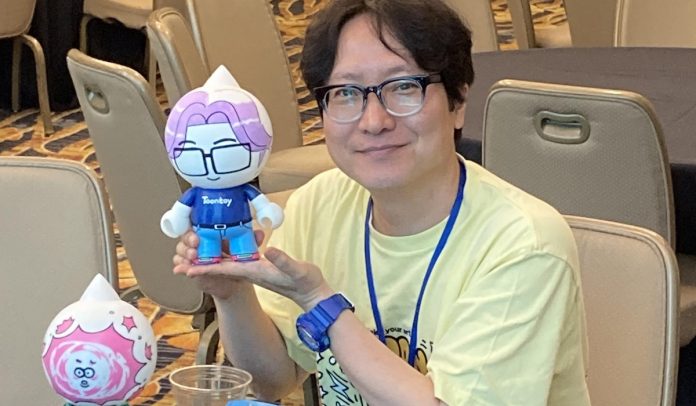 kim duckyoung featured image. a man wearing a yellow t-shirt and glasses holds a toontoy wearing glasses, a shirt and pants meant to look like him. another toontoy can be seen in the lower left.