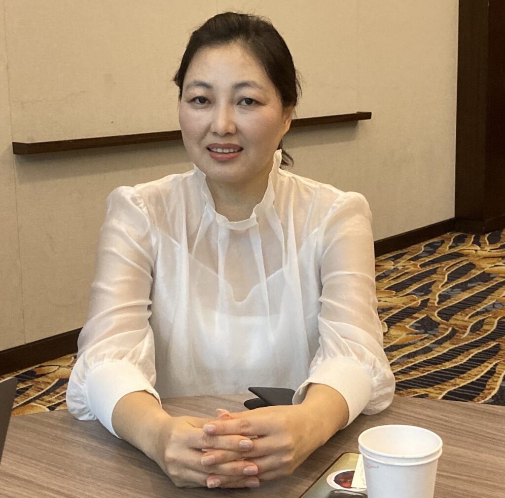 A photograph of San from KOMACON. She is wearing a white see-through blouse over a shirt. A styrofoam cup can be seen on the table.