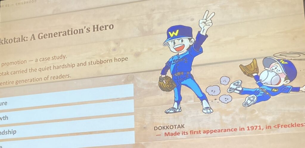 A slide reading "Dokkotak: A Generation's Hero." He made his first appearance in 1971, in "Freckles." Dokkotak carried the quiet hardship and stubborn hope of an entire generation of readers. He wears a blue baseball cap with a yellow W, and is throwing a peace sign. He can also be seen lying on the ground with a giant baseball stuffed in his mouth.